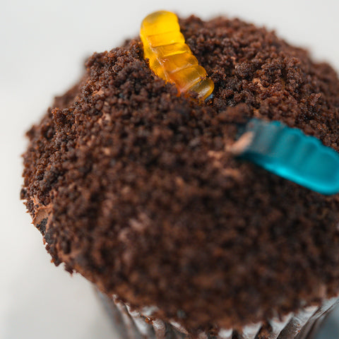 Introducing the Buffin Muffin Dirt Cake—a nostalgic treat This chocolate cupcake, inspired by the beloved chocolate muffin, is topped with crumbled chocolate cookie crumbs and finished with one yellow and one blue gummy worm for that playful “dirt with worms” look.