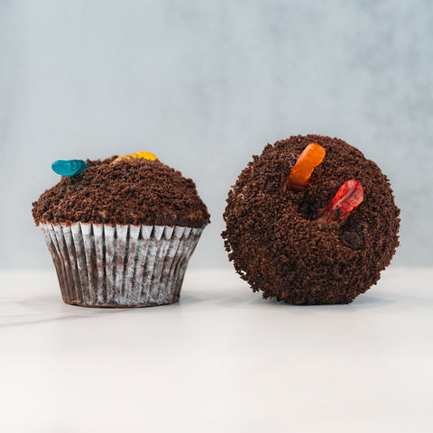Two Buffin Muffin Dirt Cake cupcakes, each resembling a decadent chocolate muffin, are topped with crumbled chocolate cookie dirt and colorful gummy worms, creating a playful garden or dirt and worms dessert look.