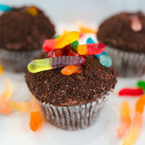 Buffin Muffin Dirt Cake: A chocolate cupcake filled with rich chocolate, topped with crumbled cookies and vibrant gummy worms, with extra gummy worms scattered on a white surface.