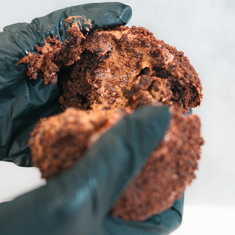 A person wearing black gloves breaks apart a Buffin Muffin Dirt Cake, revealing a gooey, rich chocolate filling at the center.