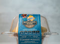 A clear plastic container holds a muffin with a blue label reading Buffalicious Baddie Blueberry Lemon Cheesecake. Nutritional info, website, and distributor details are visible on the label.