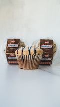 A chocolate cupcake with light icing and chocolate drizzle sits on a white surface, with two packaged CHOCOLIT EXCLUSIVE cupcakes in the background.