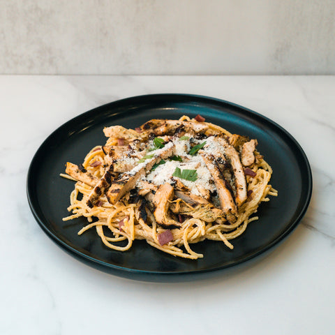 A plate of Gluten Free Carbonara Chicken Subscription features high-protein pasta with grilled chicken, cheese, herbs, and red onion on a light marble surface—a modern spin on classic Italian comfort food.