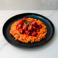 Smoky Beef Sausage and Rice: Orange jollof rice with peas, corn, and spicy smoky beef sausage cubes, garnished with green herbs on a black plate—your perfect comfort meal.