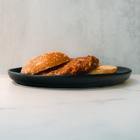 The Chicken Veggies Burger, featuring a sesame seed bun and fried chicken fillet, sits on a black plate against a light background—an ideal high-protein meal choice.