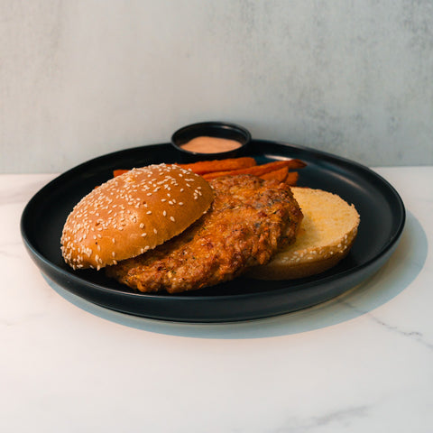 The Chicken Veggies Burger, featuring a high-protein chicken patty on a sesame seed bun, is served open-faced on a black plate with sweet potato fries and dipping sauce.
