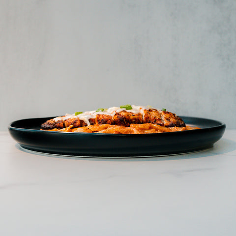 A close-up side view of Farfalle Chicken Alfredo topped with white sauce and green herbs, served on a black plate against a plain light background.