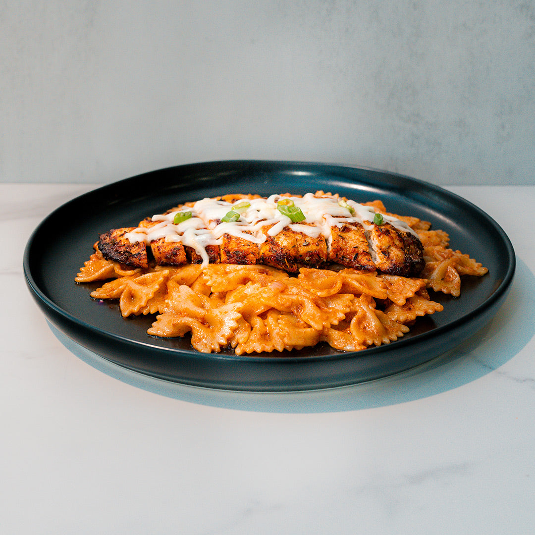Farfalle Chicken Alfredo features high-protein bowtie pasta in a creamy Alfredo sauce, topped with sliced grilled chicken breast, melted cheese, and fresh herbs—an easy ready-to-heat meal on a black plate.