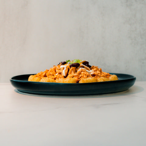 Peak Load Chick'n Tots served on a dark plate, topped with shredded cheese and beans, set on a light marble surface against a pale textured background—a high-protein comfort meal.