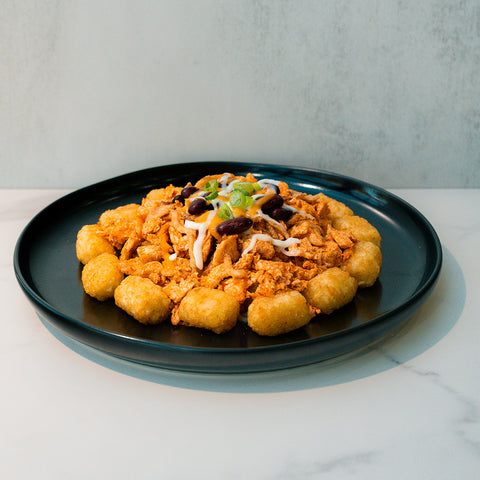 Peak Load Chick'n Tots served on a black plate: crispy tater tots in a ring with pulled chicken, beans, melted cheese, and green onions—a high-protein comfort meal on white marble.
