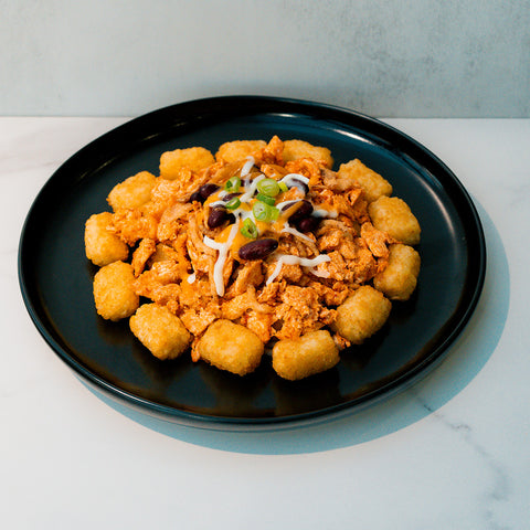 Peak Load Chick'n Tots are arranged on a black plate in a crispy ring around a center of seasoned plant-based chick'n, beans, cheese, and green onions—a tasty, high-protein comfort meal on white marble.