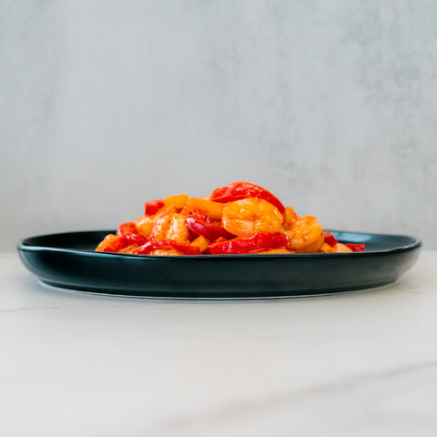 A black plate displays Hawaiian Pineapple Shrimp 1lb—a gluten-free, high-protein entrée with shrimp, red and yellow bell peppers—set on a white surface against a light gray background. Fresh flavors and vibrant colors stand out.