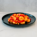 A black plate of Hawaiian Pineapple Shrimp 1lb, featuring gluten free shrimp and red bell peppers in a glossy sauce, sits on white marble with a light gray backdrop—a delectable, high-protein entrée.