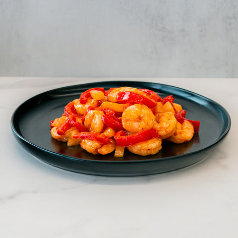 A black plate of Hawaiian Pineapple Shrimp 1lb, featuring gluten free shrimp and red bell peppers in a glossy sauce, sits on white marble with a light gray backdrop—a delectable, high-protein entrée.