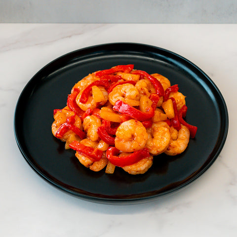 A black plate of Hawaiian Pineapple Shrimp 1lb is served with red and yellow bell pepper slices, displayed on white marble for a colorful, high-protein entrée.