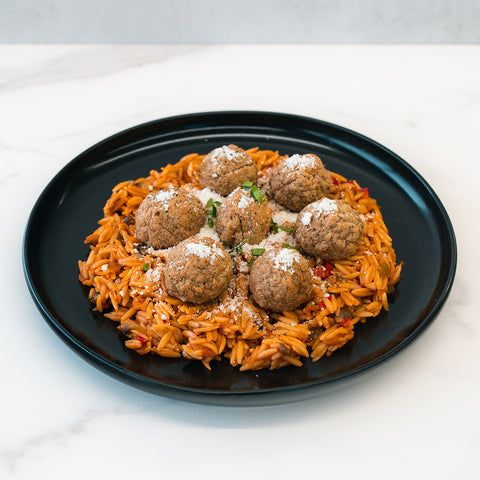 Garden Orzo Meatballs features orzo pasta in tomato sauce topped with meatballs, grated cheese, and herbs—a delicious high-protein meal prep option beautifully served on a black plate atop white marble.