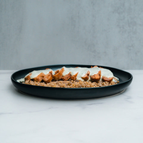 Anatolian Kebab Bulgur Rice features sliced grilled chicken breast and tender thighs with creamy white sauce, served over bulgur wheat for a Mediterranean-style meal on a dark plate against a light gray background.