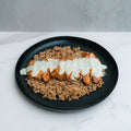 A black plate features Anatolian Kebab Bulgur Rice—sliced grilled chicken breast topped with creamy white sauce, served on bulgur wheat atop a white marble surface.