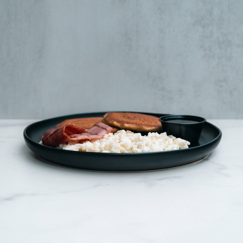 The Snickerdoodle Pancake Breakfast features scrambled eggs, ham slices, high-protein pancakes, and sugar free syrup on a dark plate set on white marble with a light gray background.