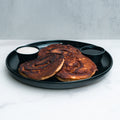 Cinnamon Roll Pancakes are served on a black plate with two small cups of white and dark sugar-free syrup, set atop white marble with a light gray background.