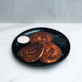 Cinnamon Roll Pancakes are served three to a plate with two sauces, one light and one dark, on a black plate set against a light gray and white marble background.