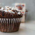 A close-up of the VIP Buffin - Rocky Road, a macro-friendly chocolate muffin topped with mini marshmallows and drizzled with chocolate sauce, set on a light surface with a blurred background.