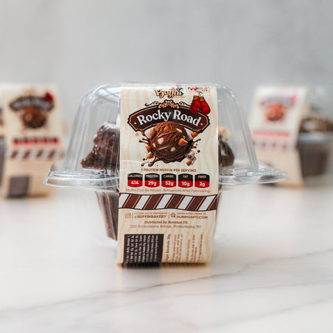 A plastic container of VIP Buffin - Rocky Road, labeled in brown and cream with nutrition facts, sits on a white surface. Two more macro-friendly snack containers are visible blurred in the background.