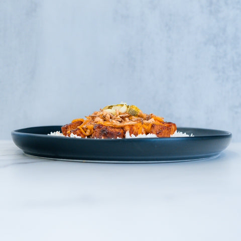 A dark plate features BBQ Meltdown—a high-protein chicken meal prep with white rice, seasoned meat, and fresh vegetable garnish—set against a light, blurred background.