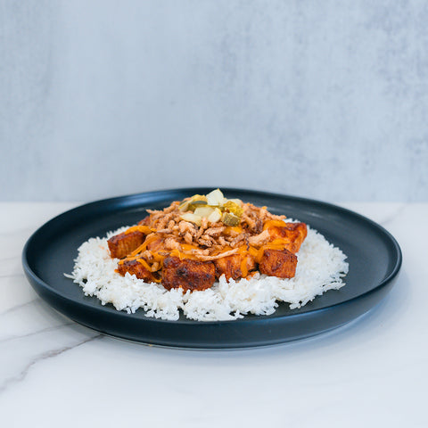 BBQ Meltdown features a black plate with white rice, fried tofu cubes, shredded cheese, ground meat, pickles, and diced onions—ideal for high-protein meal prep—presented on a marble surface against a light gray background.