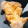 Wearing black gloves, someone pulls apart a Banana Cream Protein Dream CORE Team Edition muffin, unveiling a gooey, creamy yellow protein filling stretching between the halves.