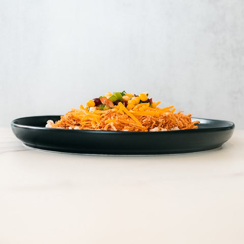 The Chicken Taco Bowl features shredded chicken, yellow cheese, corn, beans, and green onions on a black plate—a colorful, high-protein meal served on a white surface with a light grey background.
