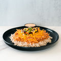 The Chicken Taco Bowl features brown rice, shredded chicken, cheddar cheese, beans, corn, and diced peppers on a black plate, with a side of creamy sauce.