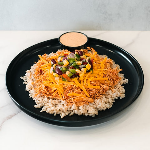 The Chicken Taco Bowl features brown rice, shredded chicken, cheddar cheese, and a colorful bean and corn salsa in the center. Served on a black plate with creamy sauce on the side, it’s a vibrant, high-protein meal.