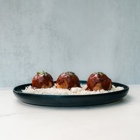 A dark plate features a high protein rice bowl topped with Caribbean Jerk Meatballs covered in sauce and herbs against a light background—Caribbean Jerk Meatballs are perfect for easy, flavorful meal prep.