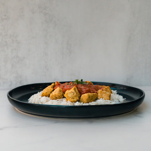 Chicken Tikka Masala: Tender golden-brown chicken in red tomato sauce and herbs over white rice on a black plate, set against a light gray background—a high-protein, gluten-free meal.