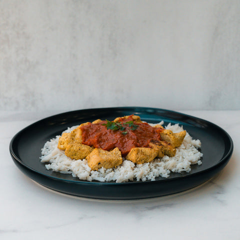 A black plate with basmati rice topped with Chicken Tikka Masala’s tender pieces in a rich tomato sauce, garnished with fresh herbs, set against a light background—this gluten-free dish is a satisfying high protein meal.