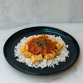 A black plate of Chicken Tikka Masala with white rice, golden tofu cubes, tomato sauce, and cilantro garnish—a high-protein, gluten-free twist on the classic dish—sits on a white surface.