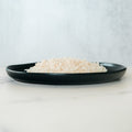 A black plate holds a mound of Competition Approved White Basmati Rice 1lb, a healthy carb source, set on a light surface with a plain pale background.