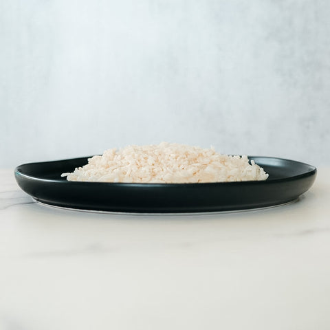 A black plate holds a mound of Competition Approved White Basmati Rice 1lb, a healthy carb source, set on a light surface with a plain pale background.