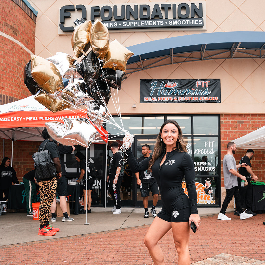 A woman in athletic wear smiles outside a nutrition store holding gold, silver, and white balloons. People gather near tents and banners promoting meals, supplements, and smoothies at the event.