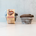 A Buffin Muffin - TwistBar chocolate caramel protein muffin in a white paper liner sits on a white surface beside a clear container with a beige TwistBar label. This high-protein treat is ideal for convenient, on-the-go snacking.