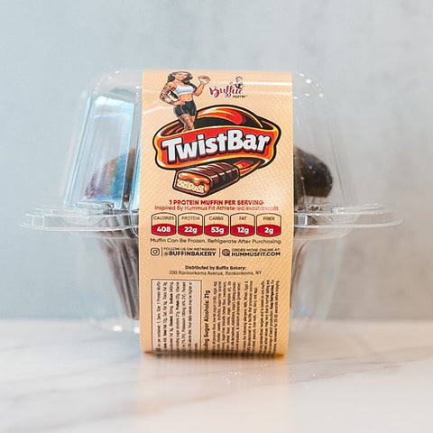 A clear-packaged Buffin Muffin - TwistBar by Buffin Bakery sits on a light surface, showing a chocolate caramel muffin. The label features nutrition details, branding, and a cartoon woman lifting weights.