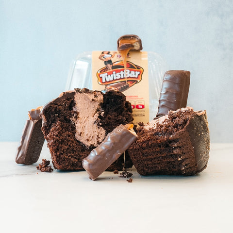 A Buffin Muffin - TwistBar protein muffin with creamy filling is sliced open, surrounded by chocolate-covered candy bars. A Buffin Muffin - TwistBar package sits in the background on a white surface against a light blue backdrop.