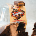 A Buffin Muffin - TwistBar protein bar with caramel filling rests on its packaging, showing a muscular cartoon woman. A high-protein chocolate muffin appears in the foreground.