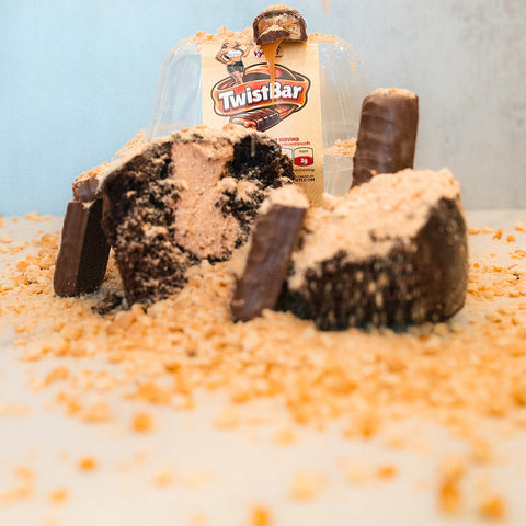 A Buffin Muffin - TwistBar, cut open to show its creamy chocolate caramel center, sits among chocolate bars and scattered crumbs. A Buffin Muffin - TwistBar wrapper appears in the background with crumbs across the surface.