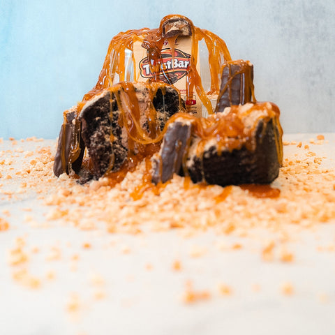 The Buffin Muffin - TwistBar is a rich chocolate caramel dessert inspired by a high-protein muffin, finished with caramel sauce and Toffee Bar pieces, all presented on white with toffee bits against a light blue backdrop.