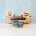 A single Buffin Muffin - TwistBar chocolate protein muffin in a white liner sits on a light surface, with two packaged Buffin Muffins blurred in the background against a light wall.