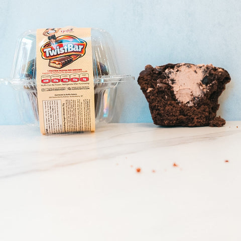 A Buffin Muffin - TwistBar, a chocolate caramel dessert in a clear plastic container, sits next to a high protein chocolate muffin with creamy filling on a white surface with a light blue background.