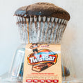 A Buffin Muffin - TwistBar with a domed top is upside down on a clear plastic container, partially covering its candy wrapper, which displays the logo, nutrition facts, and an illustration of a woman running.