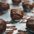 Close-up of 3 Banana Chocolate Protein Poppers on a light surface, each coated in glossy chocolate drizzle. The focus is on the center popper, showing its smooth finish—a delicious and healthy snack option.